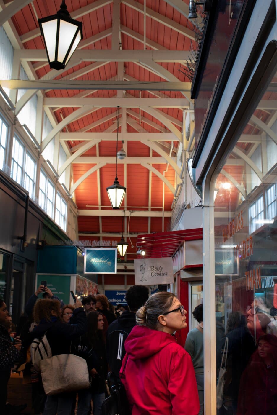 A covered market under a red timber roof, shoppers below and a woman in a red coat