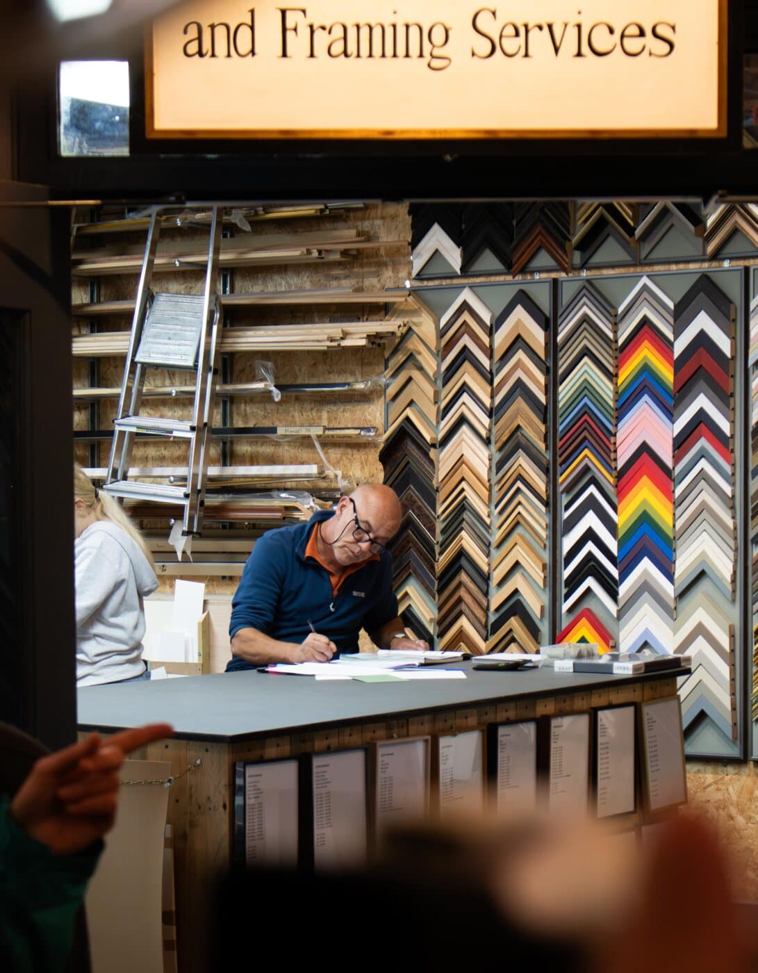 A framer at his counter under an “and Framing Services” sign, walls of frame samples behind