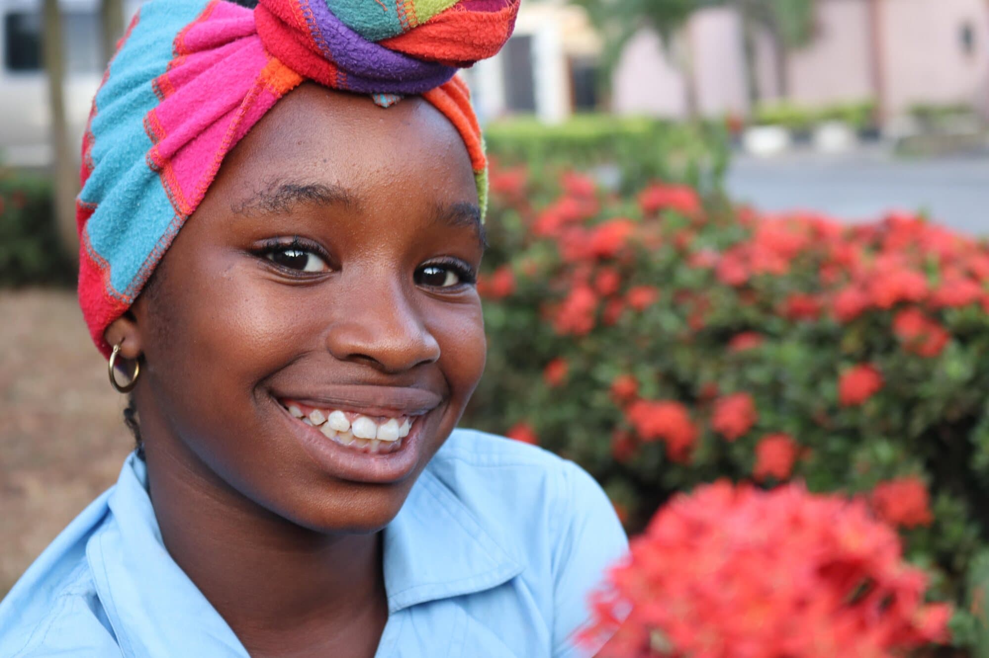 A smiling young woman in a bright striped headwrap, red flowers behind her